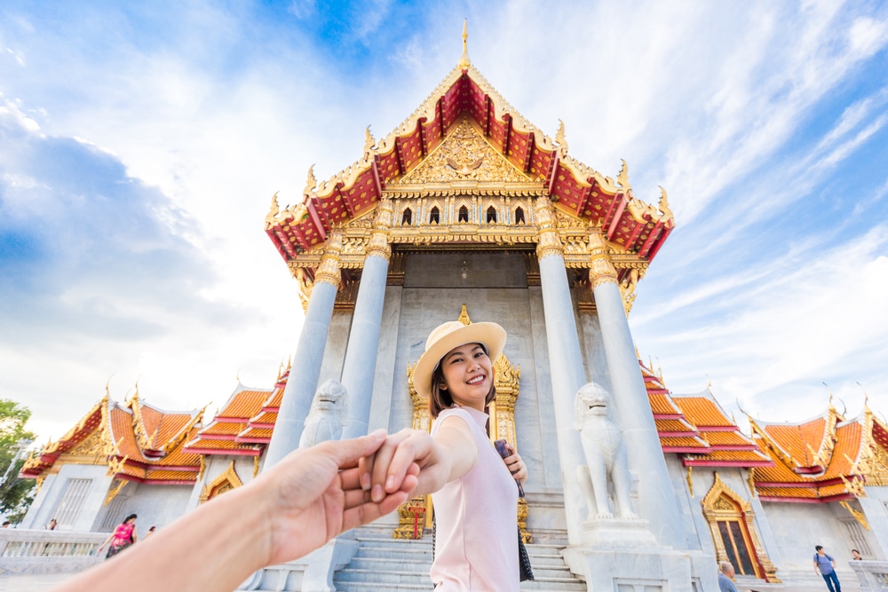 บุคคลยืนยิ้มและจับมืออีกฝ่าย โดยมีวิหารอันวิจิตรงดงามซึ่งมีรายละเอียดสีแดงและสีทอง ที่เดทในกรุงเทพกลางคืน และมีเสาเป็นพื้นหลังใต้ท้องฟ้าสีครามและมีเมฆกระจัดกระจาย เหมาะสำหรับการเดทในกรุงเทพกลางคืน