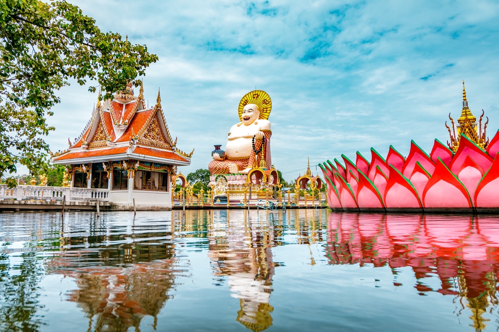 กลุ่มวัดริมน้ำ มีพระพุทธรูปขนาดใหญ่ สถานที่ท่องเที่ยวสุราษ โครงสร้างดอกบัวสีสันสดใส สถาปัตยกรรมไทยแบบดั้งเดิม และต้นไม้สีเขียวหนาแน่นตัดกับท้องฟ้าสีคราม