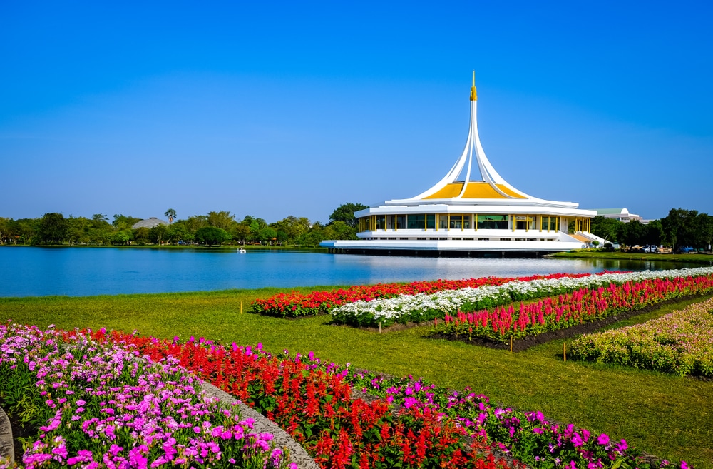 ศาลาสีขาวเหลืองริมทะเลสาบในสวนสาธารณะ โดยมีสวนดอกไม้หลากสีสันอยู่เบื้องหน้าและท้องฟ้าสีครามเป็นพื้นหลัง ทำให้เกิดบรรยากาศอันน่าหลงใหล ที่เดทในกรุงเทพกลางคืน ที่เที่ยวในกรุงเทพธรรมชาติ