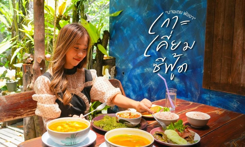 ร้านอาหารพังงา ผู้หญิงคนหนึ่งนั่งอยู่ที่โต๊ะไม้ด้านนอก เลือกอาหารจากชามและจานต่างๆ หลายจาน โดยมีผนังสีฟ้าและพื้นหลังเขียวขจีเป็นฉากหลัง ร้านอาหารเมืองพังงาเขียนด้วยข้อความภาษาไทยทางด้านขวา