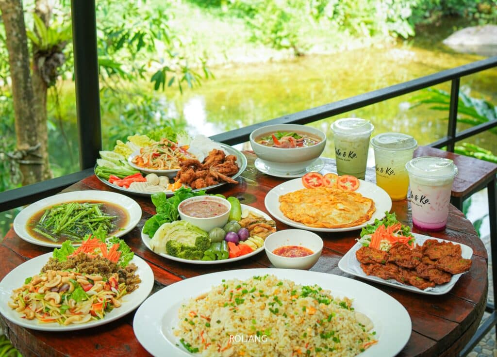 โต๊ะที่เต็มไปด้วยอาหารไทยนานาชนิด รวมถึงผักผัด สลัด เนื้อทอด และเครื่องดื่ม ตั้งอยู่กลางแจ้งอันเขียวชอุ่มริมน้ำ หนึ่งใน ที่เที่ยวพังงา ประสบการณ์อันน่าหลงใหลมากมายที่คุณจะพบได้เมื่อมาเยือนพังงา