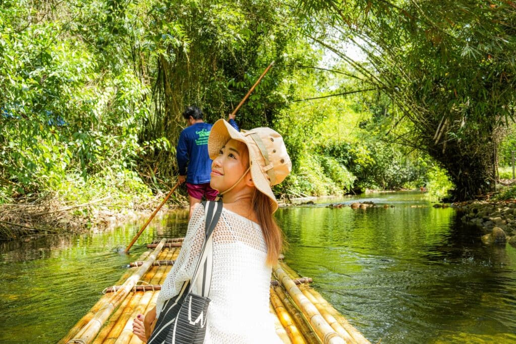 ผู้หญิงสวมหมวกและเสื้อสีขาวกำลังนั่งอยู่บนแพไม้ไผ่ในแม่น้ำที่ล้อมรอบด้วยแมกไม้เขียวขจีใน พังงาที่เที่ยว โดยมีผู้ยืนบังคับแพอยู่ด้านหลัง