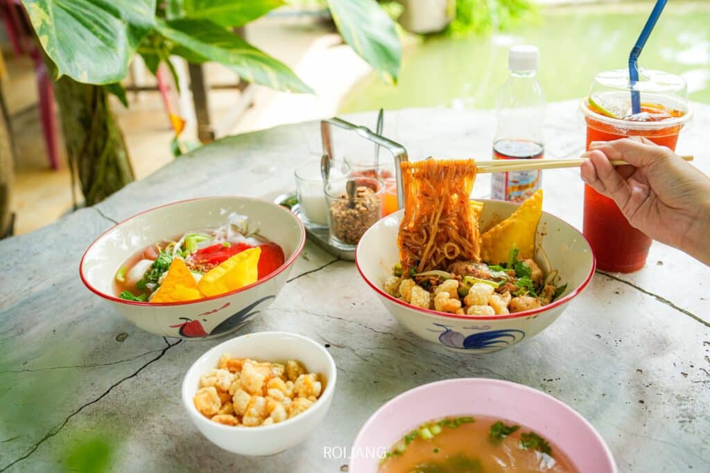 โต๊ะที่ร้านเขาหลักประกอบด้วยชามซุปก๋วยเตี๋ยวต่างๆ ชามเล็กๆ ที่ใส่ท็อปปิ้งทอด เครื่องดื่มสีแดงหนึ่งแก้วพร้อมหลอด ขวดน้ำหนึ่งขวด และมือถือตะเกียบถือบะหมี่ ร้านอาหารเขาหลัก