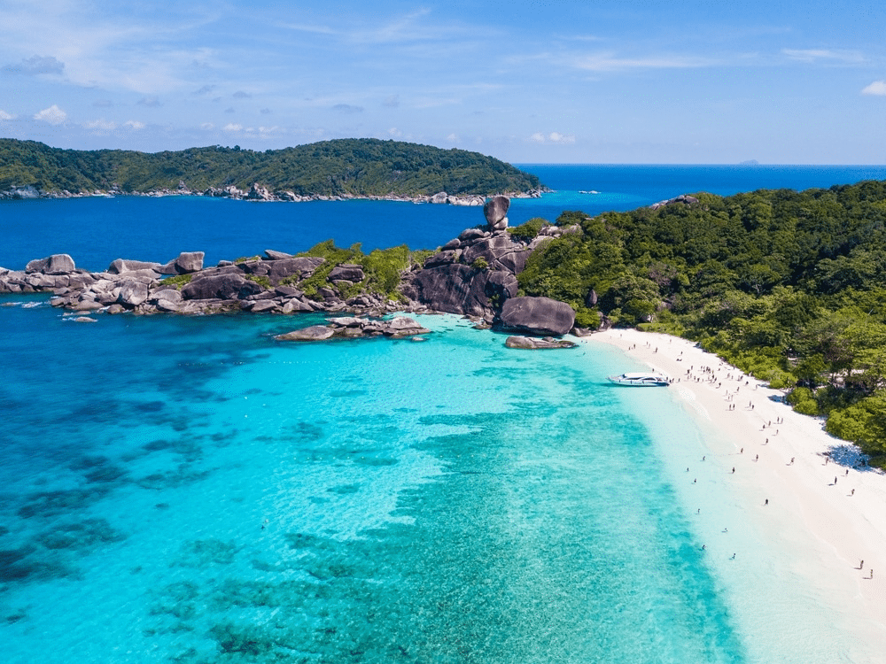 ทิวทัศน์อันงดงามของชายหาดสีฟ้าครามที่มีหาดทรายสีขาว ที่เที่ยวเดือนมีนาคม ล้อมรอบด้วยเนินเขาสีเขียวชอุ่มและแนวหิน ภายใต้ท้องฟ้าสีครามสดใส เป็นที่เที่ยวที่ลงตัวสุดๆ โดยมีผู้คนกระจัดกระจายไปตามแนวชายฝั่ง