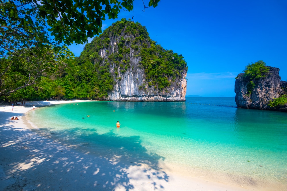 ชายหาดเขตร้อนที่มีหาดทรายสีขาว น้ำทะเลสีฟ้าครามใส และหน้าผาสีเขียวชอุ่มภายใต้ท้องฟ้าสีฟ้าสดใสบนเกาะในประเทศไทยในประเทศไทย ผู้คนกำลังว่ายน้ำและพักผ่อนบนฝั่ง เกาะที่สวยที่สุดในประเทศไทย