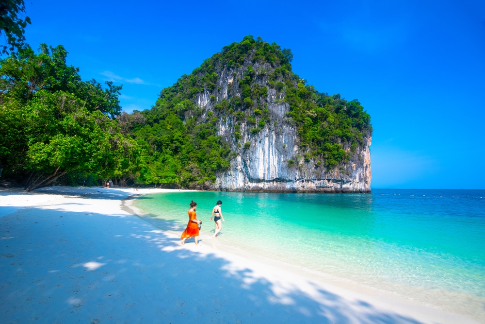 คนสองคนกำลังเดินไปตามชายหาดอันบริสุทธิ์ที่มีหาดทรายสีขาวและน้ำทะเลสีฟ้าครามใส บนเกาะในประเทศไทยในประเทศไทย เนินเขาหินเขียวชอุ่มเป็นฉากหลังภายใต้ท้องฟ้าสีฟ้าสดใส เกาะที่สวยที่สุดในประเทศไทย