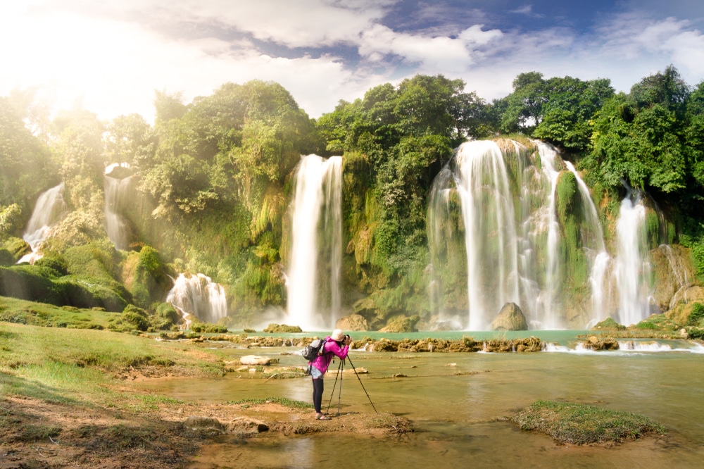 คนในชุดสีชมพูยืนด้วยขาตั้งกล้องและกล้อง น้ำตกที่สวยที่สุดในโลก  เพื่อถ่ายภาพน้ำตกสวยที่สุดในโลกในขณะที่น้ำตกขนาดใหญ่ไหลลงสู่แอ่งน้ำอันเงียบสงบที่ล้อมรอบด้วยต้นไม้เขียวขจีภายใต้ท้องฟ้าที่มีเมฆบางส่วน