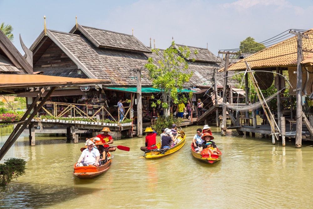 นักพายเรือในเรือคายัคแบบดั้งเดิมสีสันสดใสล่องไปตามลำคลองระหว่างอาคารไม้ทรงไทยที่มีหลังคามุงจากในวันที่อากาศแจ่มใส มองเห็นผู้คนเดินอยู่บนท่าเรือไม้เป็นฉากหลัง ทำให้จุดที่มีเสน่ห์แห่งนี้เหมาะสำหรับการเที่ยวหน้าโรแมนติกฝน เที่ยวเมืองรอง