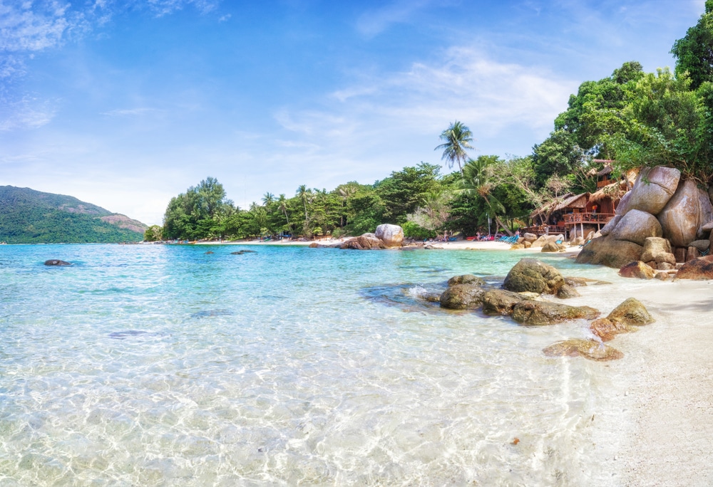 ชายหาดที่มีน้ำทะเลสีฟ้าครามใส ทรายขาว หิน ต้นปาล์ม และทิวทัศน์อันไกลโพ้นของโครงสร้างไม้ภายใต้ท้องฟ้าสีครามในวันที่แดดจ้า บรรยากาศอันงดงามนี้สามารถเป็นส่วนหนึ่งของเกาะได้อย่างง่ายดายในประเทศไทย เกาะที่สวยที่สุดในประเทศไทย