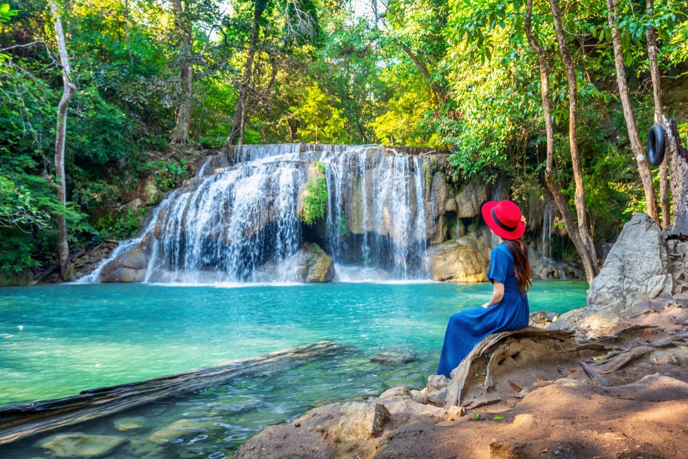 คนที่สวมหมวกสีแดงและชุดสีน้ำเงินนั่งข้างน้ำตกสีฟ้าครามสดใส น้ำตกที่สวยที่สุดในประเทศไทย น้ำตกในประเทศนี้ ล้อมรอบด้วยต้นไม้สีเขียวชอุ่ม