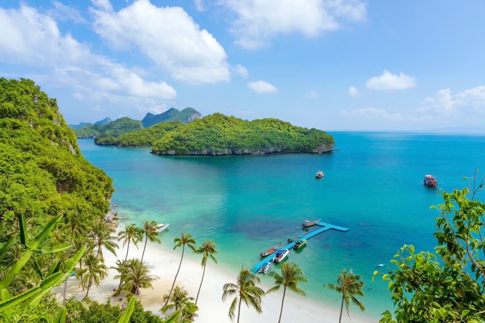 ชายหาดเขตร้อนที่มีหาดทรายขาว ต้นปาล์ม น้ำทะเลสีฟ้าคราม และเรือจอดอยู่ที่ท่าเรือ มองเห็นเกาะสีเขียวเป็นพื้นหลังภายใต้ท้องฟ้าที่มีเมฆบางส่วน สะท้อนถึงแก่นแท้ของเกาะในประเทศไทยในประเทศไทย เกาะที่สวยที่สุดในประเทศไทย