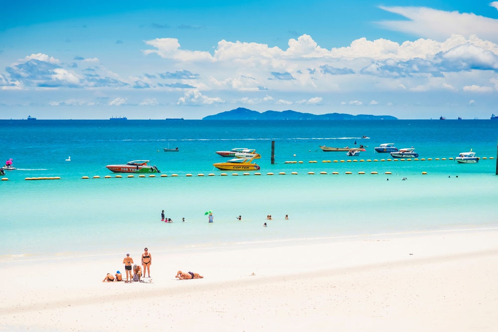 ฉากชายหาดที่มีแสงแดดสดใสซึ่งมีผู้คนกำลังอาบแดดบนหาดทรายสีขาว เรือที่ลอยอยู่ในน้ำทะเลสีฟ้าคราม และเกาะต่างๆ ในประเทศไทยที่มองเห็นได้ในพื้นหลังภายใต้ท้องฟ้าที่มีเมฆบางส่วน เกาะที่สวยที่สุดในประเทศไทย