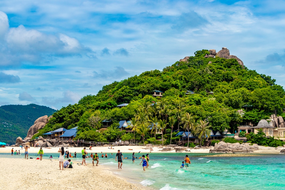 ผู้คนเพลิดเพลินกับชายหาดและน้ำทะเลสีฟ้าครามของเกาะเขตร้อน เกาะแห่งนี้ในประเทศไทย โดยมีเนินเขาสีเขียวชอุ่มภายใต้ท้องฟ้าที่มีเมฆบางส่วน เกาะที่สวยที่สุดในประเทศไทย