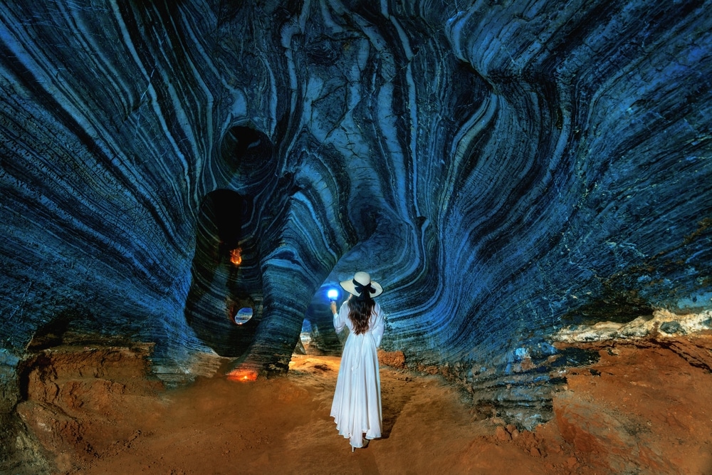 ผู้หญิงในชุดขาวและหมวกยืนอยู่ในถ้ำสีฟ้าขนาดใหญ่ ถือโคมไฟและมองดูผนังถ้ำ ที่เที่ยวแม่สอด