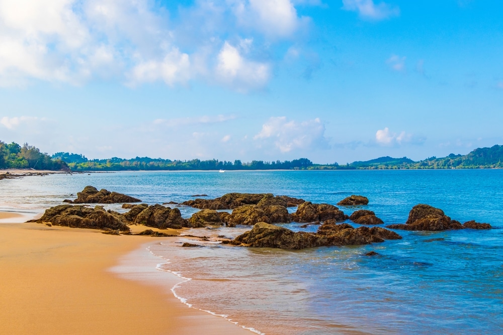 หาดทรายที่มีหินกระจัดกระจายทอดไปสู่มหาสมุทรสีฟ้าอันเงียบสงบภายใต้ท้องฟ้าที่มีเมฆบางส่วน ชวนให้นึกถึงเกาะในประเทศไทยในประเทศไทย มองเห็นต้นไม้และเนินเขาที่อยู่ห่างไกลเป็นฉากหลัง เกาะที่สวยที่สุดในประเทศไทย