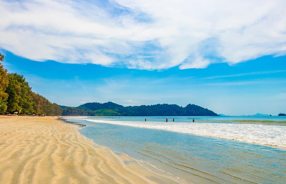 หาดทรายที่มีคลื่นอ่อนโยน ท้องฟ้าสีฟ้าใส และร่างที่ห่างไกลสองสามตัวกำลังลุยน้ำบนเกาะในประเทศไทยในประเทศไทย เนินเขาที่ปกคลุมไปด้วยป่าไม้มองเห็นได้เป็นฉากหลัง เกาะที่สวยที่สุดในประเทศไทย