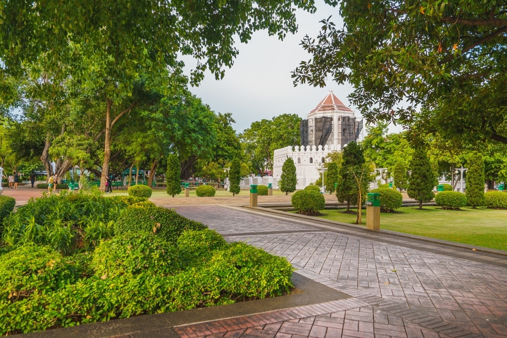 ทางเดินปูผ่านสวนสาธารณะที่มีต้นไม้เขียวขจี นำไปสู่อาคารหินสีขาวที่มี ที่เดินเล่นกทม หอคอยหลังคาสีแดงเป็นพื้นหลัง เป็นสถานที่อันเงียบสงบสำหรับเดินเล่นกทม.