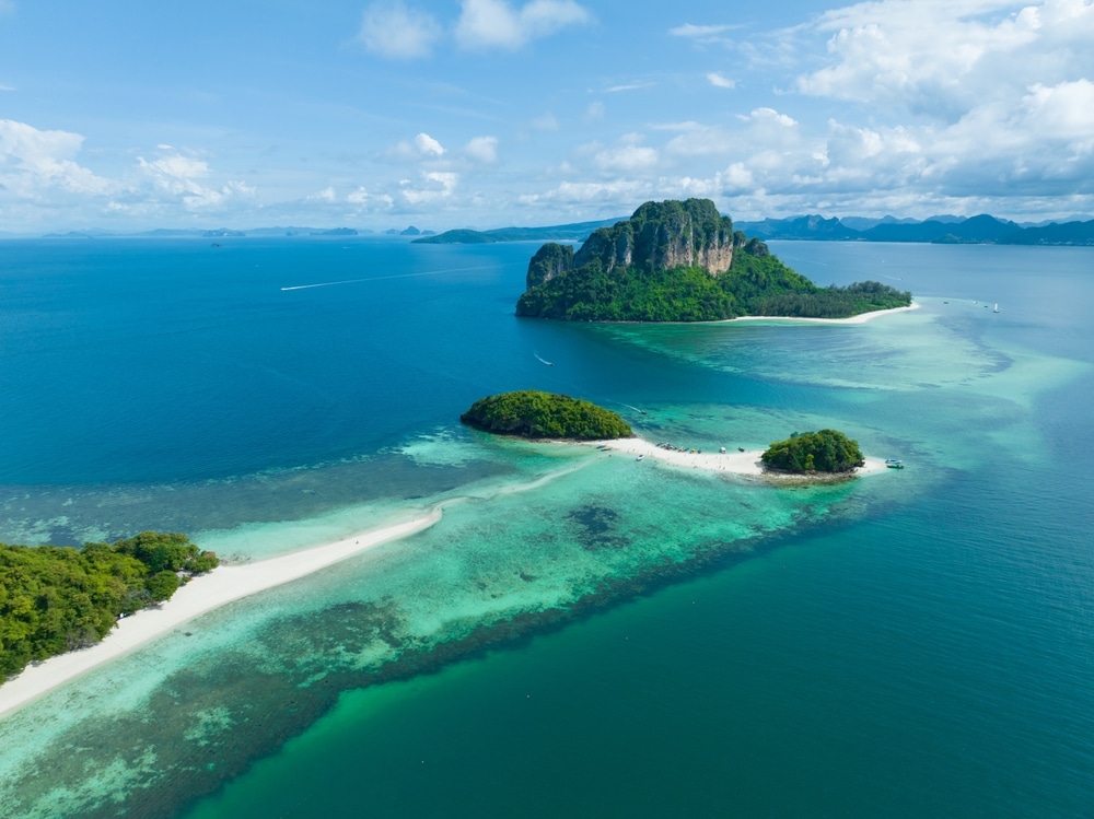 มุมมองทางอากาศของเกาะในประเทศไทยในประเทศไทยที่มีพืชพรรณสีเขียว หาดทรายสีขาว และน้ำทะเลสีฟ้าคราม มีหน้าผาหินปูนสูงเป็นพื้นหลังภายใต้ท้องฟ้าสีครามและมีเมฆกระจัดกระจาย เกาะที่สวยที่สุดในประเทศไทย 