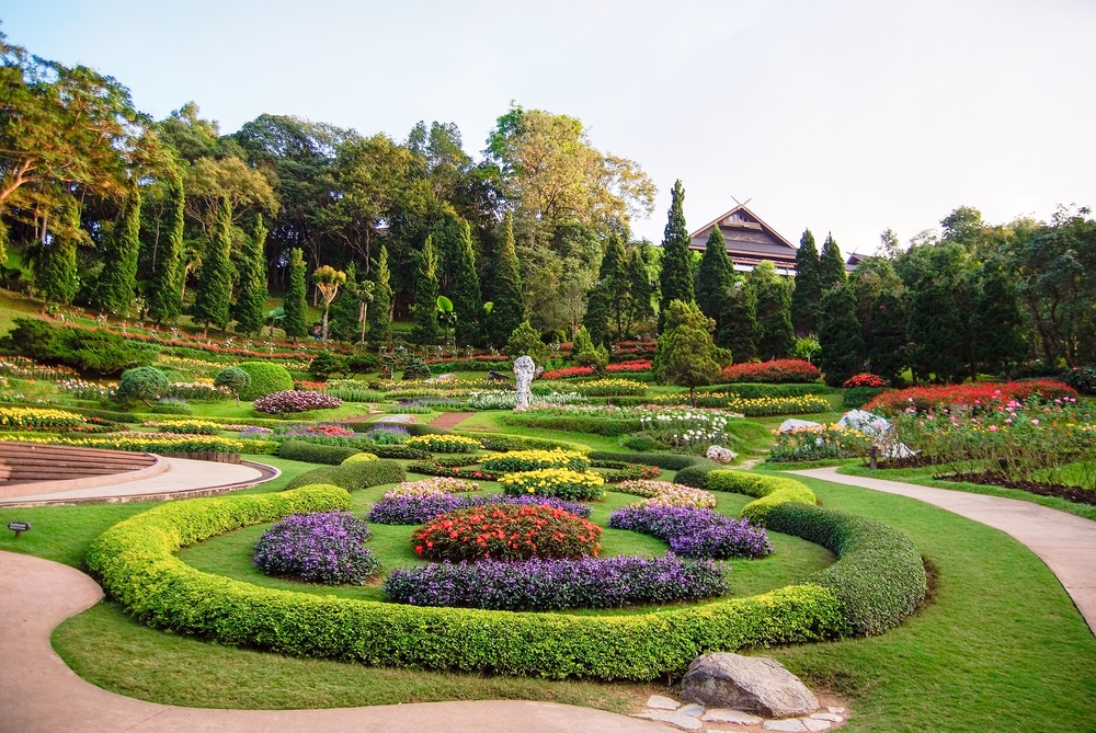 สวนภูมิทัศน์ที่มีแปลงดอกไม้สีสันสดใส พุ่มไม้สีเขียว ทางเดินคดเคี้ยว และรูปปั้นตรงกลาง มองเห็นอาคารไม้แบบดั้งเดิมเป็นพื้นหลังที่รายล้อมไปด้วยต้นไม้สูง เหมาะสำหรับผู้ที่ต้องการท่องเที่ยวในช่วงฤดูร้อนภาคเหนือ เที่ยวฤดูร้อนภาคเหนือ