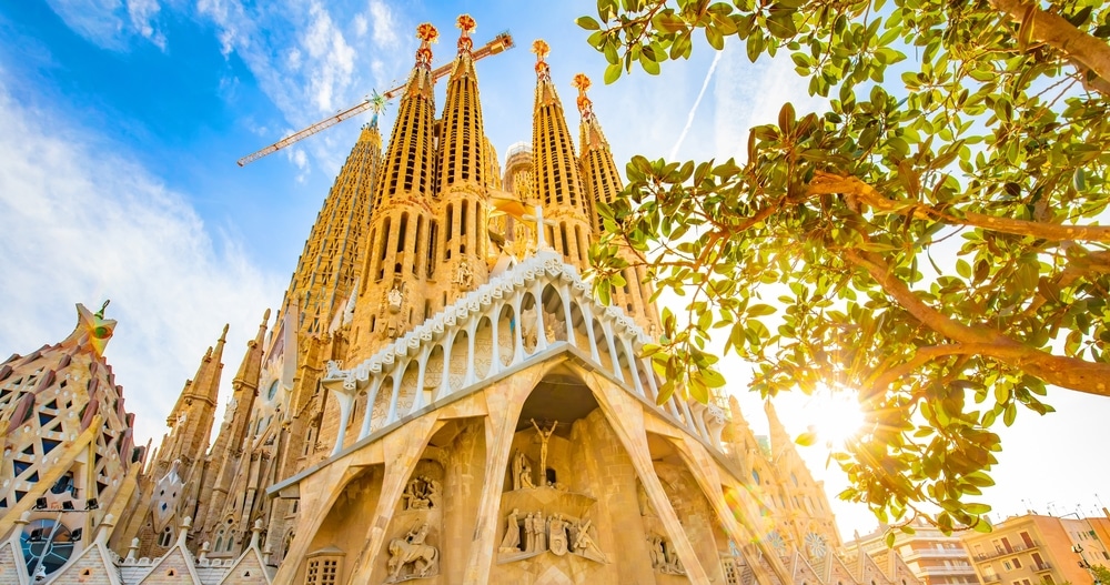 มุมมองของ Sagrada Familia ในบาร์เซโลนา ประเทศสเปน เที่ยวต่างประเทศงบน้อย มียอดแหลมที่ซับซ้อนและเครนก่อสร้างที่มองเห็นได้ ล้อมรอบด้วยต้นไม้และแสงแดดจ้า ฉากนี้ชวนให้นึกถึงความอัศจรรย์ราวกับได้ค้นพบร้านอาหารพัทยาท่ามกลางสถานที่ท่องเที่ยวอันคึกคักของชลบุรี