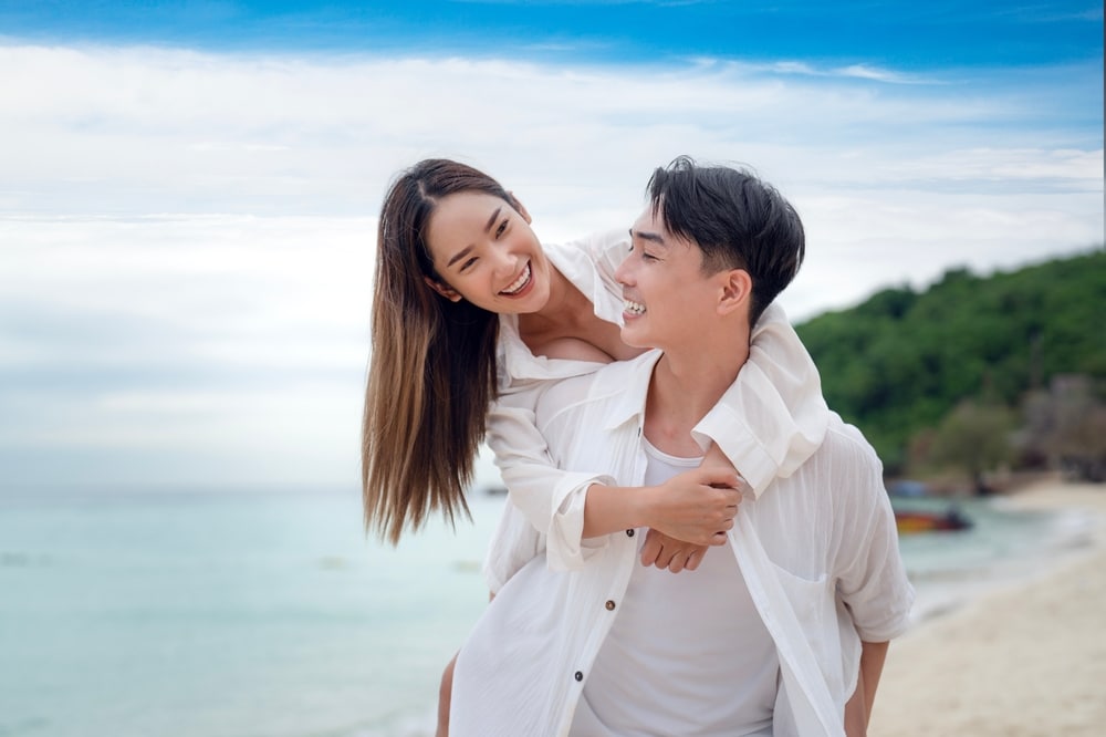 ผู้ชายขี่หลังผู้หญิงบนชายหาดในพัทยา ทั้งคู่ยิ้มและสวมเสื้อเชิ้ตสีขาว โดยมีทะเลและเนินเขาสีเขียวเป็นฉากหลัง