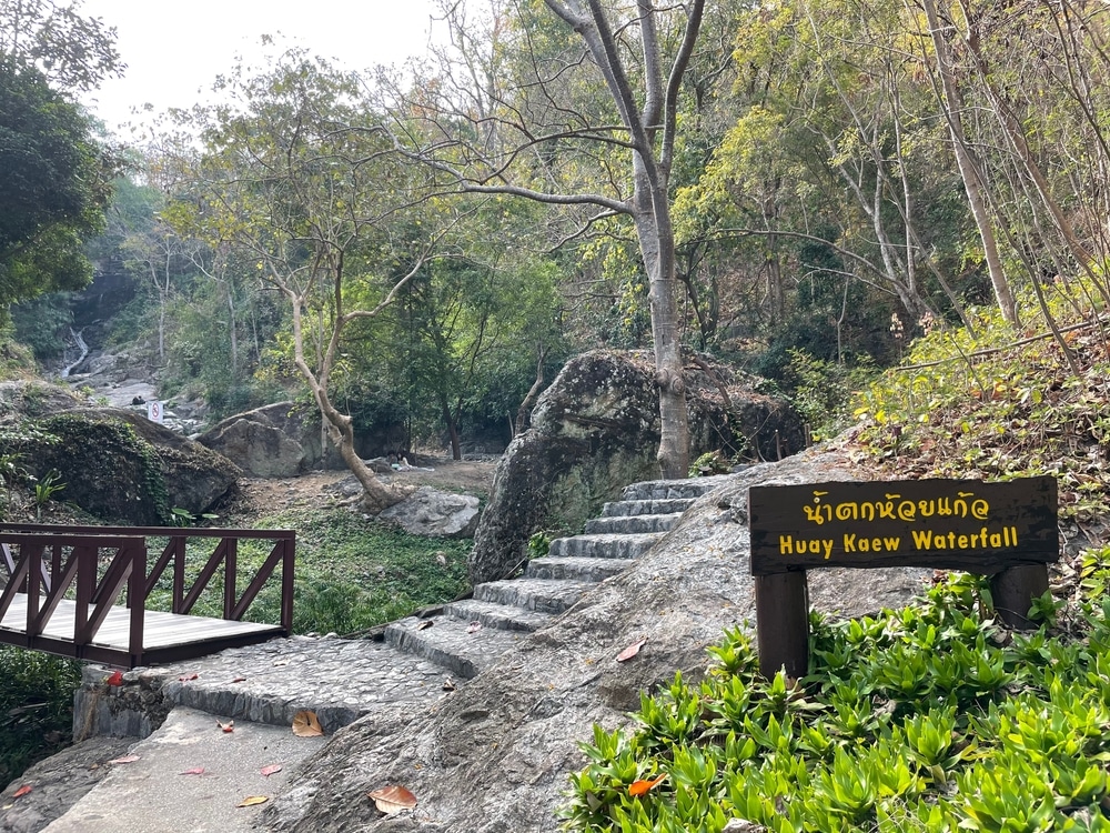 ขั้นบันไดหินและสะพานไม้เล็กๆ ทอดผ่านพื้นที่ป่าไปสู่น้ำตก โดยมีป้ายเขียนว่า "น้ำตกห้วยแก้ว" เป็นภาษาอังกฤษและภาษาไทย เที่ยวภาคเหนือช่วงฤดูร้อนเหมาะสำหรับผู้รักธรรมชาติที่แสวงหาความสงบและความสวยงาม เที่ยวภาคเหนือฤดูร้อน