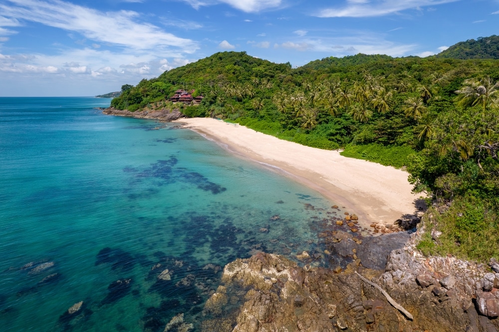 ชายหาดเขตร้อนอันเงียบสงบที่มีน้ำทะเลสีฟ้าครามใส หาดทราย และพืชพันธุ์สีเขียวชอุ่มบนเนินเขาโดยรอบภายใต้ท้องฟ้าสีครามที่มีเมฆบางส่วน ซึ่งถือเป็นศูนย์รวมของเกาะในประเทศไทยในประเทศไทย เกาะที่สวยที่สุดในประเทศไทย