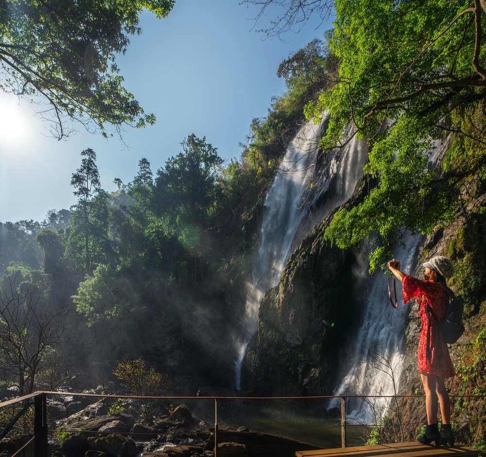 คนในชุดสีแดงกำลังถ่ายรูปน้ำตกใน น้ำตกที่สวยที่สุดในประเทศไทย ประเทศไทยที่ลดหลั่นลงมาตามหน้าผาหินที่ล้อมรอบด้วยป่าเขียวขจีในวันที่แดดจ้า