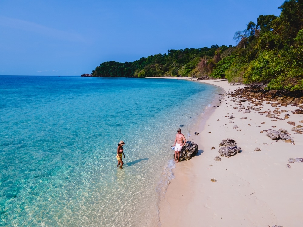 คนหนึ่งลุยน้ำสีฟ้าใส ขณะที่อีกคนหนึ่งยืนอยู่บนโขดหินบนชายหาดเขตร้อน ล้อมรอบด้วยต้นไม้เขียวขจี ชวนให้นึกถึงการอยู่บนเกาะในประเทศไทยในประเทศไทย เกาะที่สวยที่สุดในประเทศไทย