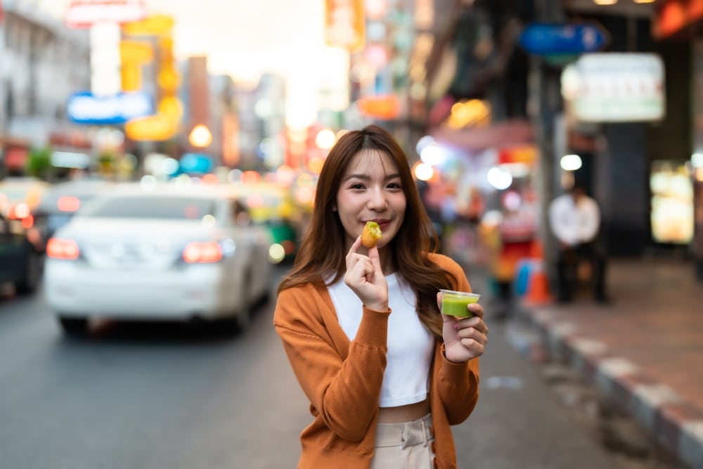 หญิงสาวกำลังเพลิดเพลินกับอาหารข้างทางบนถนนในเมืองที่วุ่นวายในกทม โดยถือไม้เสียบในมือข้างหนึ่งและอีกมือถือถ้วยซอส โดยมีป้ายไฟสว่างจ้าและรถที่วิ่งพลุกพล่านเป็นฉากหลัง