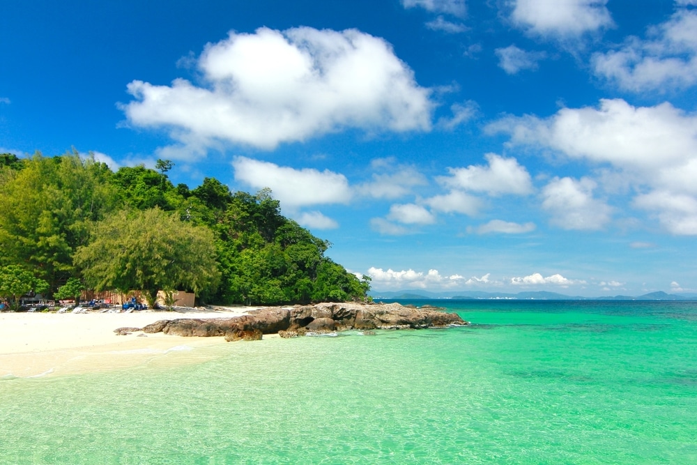 ชายหาดเขตร้อนบนเกาะแห่งนี้ในประเทศไทยมีน้ำทะเลสีฟ้าครามใส หาดทรายขาวทอดยาว และพืชพันธุ์สีเขียวชอุ่มภายใต้ท้องฟ้าสีครามสดใสและมีเมฆกระจายอยู่ เกาะที่สวยที่สุดในประเทศไทย