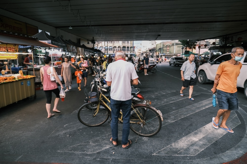 ตลาดนัดริมถนนอันคึกคักใต้สะพานลอยขนาดใหญ่ใน ที่เดินเล่นกทม โดยมีผู้คนเดินและชายยืนอยู่ข้างจักรยาน แผงลอยของผู้ขายและไฟแขวนมองเห็นได้ในพื้นหลัง