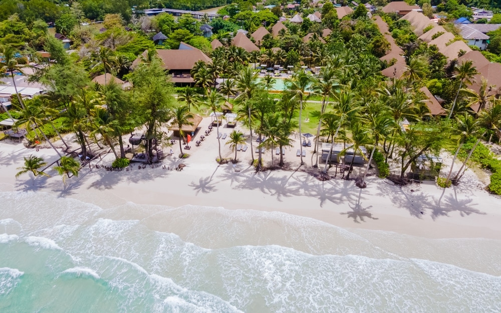 วิวมุมสูงของรีสอร์ทริมชายหาดที่มีต้นปาล์ม เก้าอี้นั่งเล่น และสระว่ายน้ำ ล้อมรอบด้วยแมกไม้เขียวขจีและติดกับมหาสมุทร เหมาะสำหรับการพักผ่อนที่เที่ยวหน้าฝนโรแมนติก เที่ยวเมืองรอง