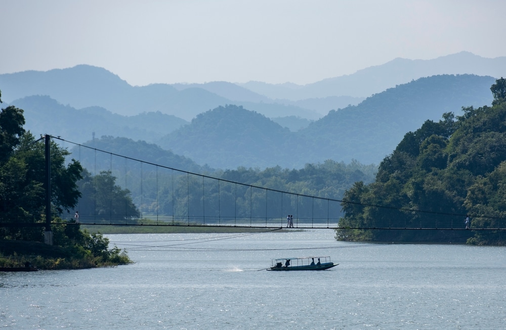 เรือแล่นไปในแม่น้ำกว้างใหญ่โดยมีสะพานแขวนคนเดินและภูเขาที่ปกคลุมด้วยป่าหมอกเป็นฉากหลัง เหมาะสำหรับการพักผ่อนแบบครอบครัว พาลูกเที่ยวธรรมชาติใกล้กรุงเทพ