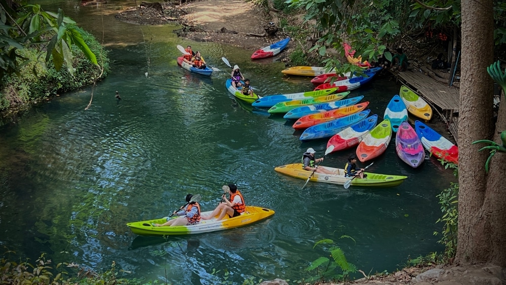 นักพายเรือคายัคกลุ่มหนึ่งพายเรือไปตามแม่น้ำอันเงียบสงบที่รายล้อมไปด้วยป่าไม้ระหว่างที่เที่ยวในการผจญภัย โดยมีเรือคายัคหลากสีสันเรียงรายไปตามริมน้ำ ที่เที่ยวเดือนมีนาคม