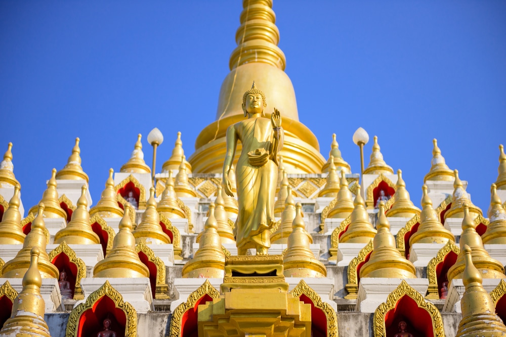 รูปปั้นพระพุทธรูปยืนสีทองล้อมรอบด้วยยอดแหลมเล็กๆ สีทอง ตั้งตัดกับท้องฟ้าสีฟ้าใส ที่เที่ยวแม่สอด