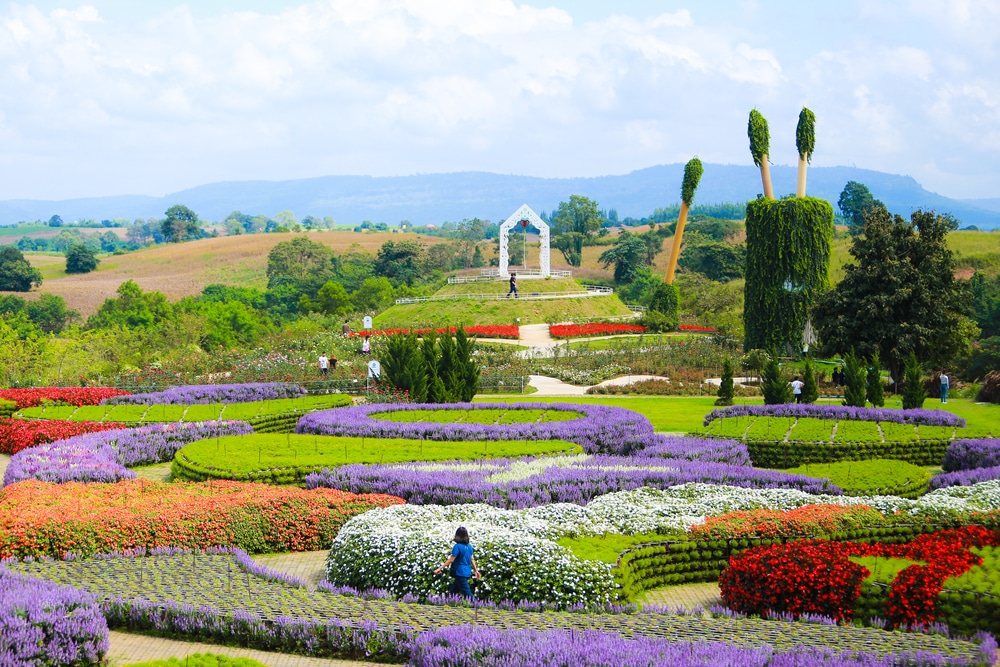 สวนสวยพร้อมแปลงดอกไม้หลากสีสัน ทางเดินคดเคี้ยว โครงสร้างโค้งสีขาว และเนินเขาอันห่างไกลภายใต้ท้องฟ้าสีครามและมีเมฆ มอบสถานที่ท่องเที่ยวหน้าฝนโรแมนติกในอุดมคติ