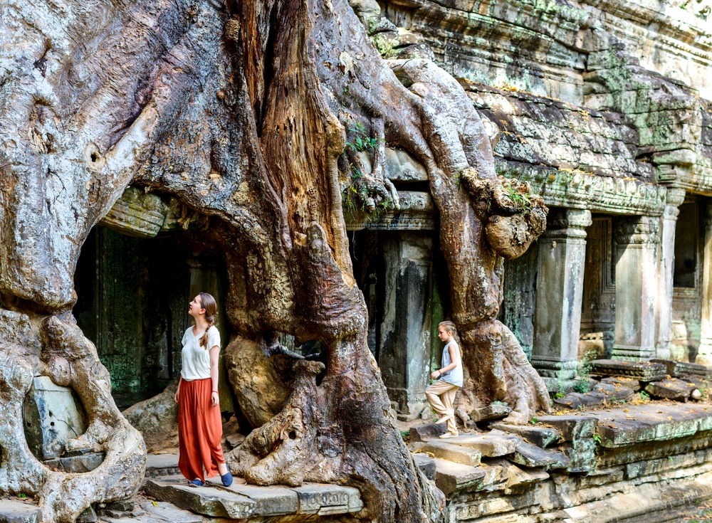 คนสองคนสำรวจวัดโบราณแห่งหนึ่งในกัมพูชาซึ่งมีรากต้นไม้ ท่องเที่ยวกัมพูชา ขนาดใหญ่เติบโตอยู่เหนือโครงสร้างของวัด คนหนึ่งยืนอยู่ใต้ราก ส่วนอีกคนหนึ่งเดินอยู่ใกล้ๆ วัดมีเสาหินและผนังที่ผุกร่อน สะท้อนถึงประวัติศาสตร์อันยาวนานของท่องเที่ยวกระเป๋าเดินทาง