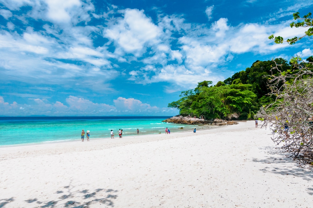 หาดทรายที่มีน้ำทะเลสีฟ้าครามใส ผู้คนเดินไปตามชายฝั่ง และต้นไม้เขียวชอุ่มเป็นพื้นหลังภายใต้ท้องฟ้าที่มีเมฆบางส่วนบนเกาะในประเทศไทยในประเทศไทย เกาะที่สวยที่สุดในประเทศไทย