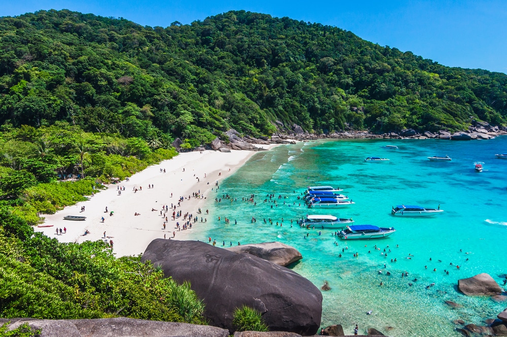 ชายหาดที่มีหาดทรายขาวและน้ำทะเลสีฟ้าครามใส ล้อมรอบด้วยเนินเขาสีเขียวชอุ่มบนเกาะในประเทศไทยในประเทศไทย เรือจอดทอดสมออยู่ใกล้ชายฝั่งในขณะที่ผู้คนว่ายน้ำและพักผ่อนบนชายหาด เกาะที่สวยที่สุดในประเทศไทย