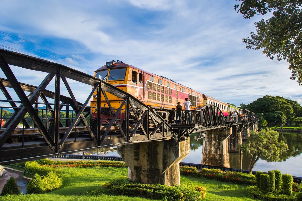 รถไฟข้ามสะพานเหล็กข้ามแม่น้ำที่มีต้นไม้และแมกไม้เขียวขจีในวันที่อากาศแจ่มใส จัดแสดงที่เที่ยวหน้าฝนโรแมนติก ที่เที่ยวหน้าฝนโรแมนติก