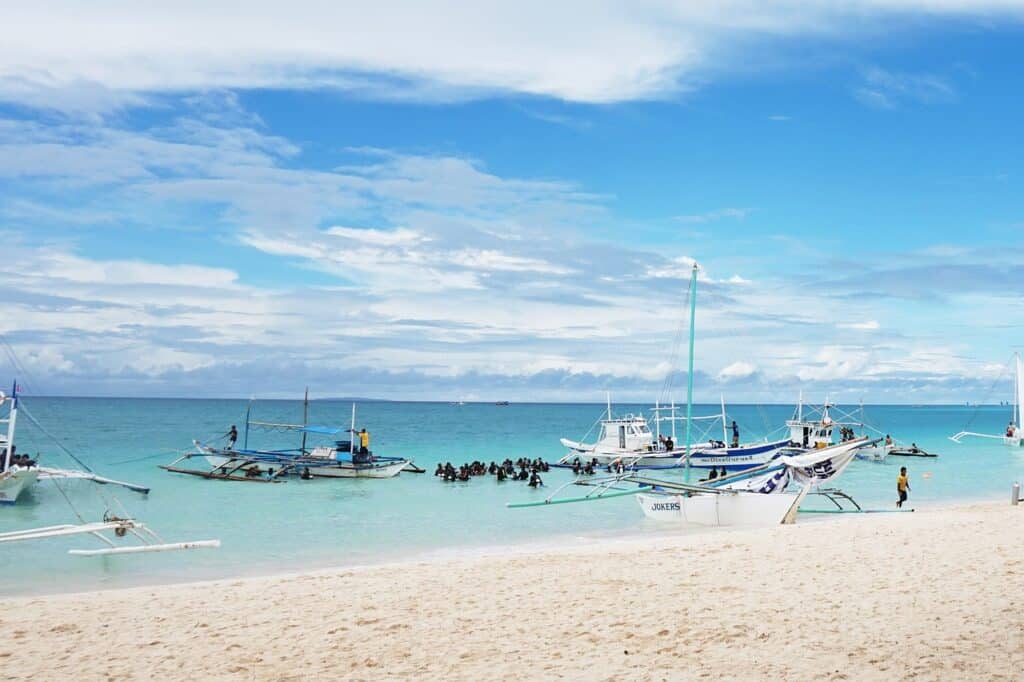 ชายหาดทรายมีเรือหลายลำจอด ฮันนีมูน ที่ไหนดี ทอดสมออยู่ในน้ำทะเลสีฟ้าใส มีกลุ่มคนสวมชุดดำน้ำสีดำอยู่ใกล้ๆ เรือ ท้องฟ้ามีเมฆบางส่วนและมีจุดสีฟ้าเป็นหย่อมๆ