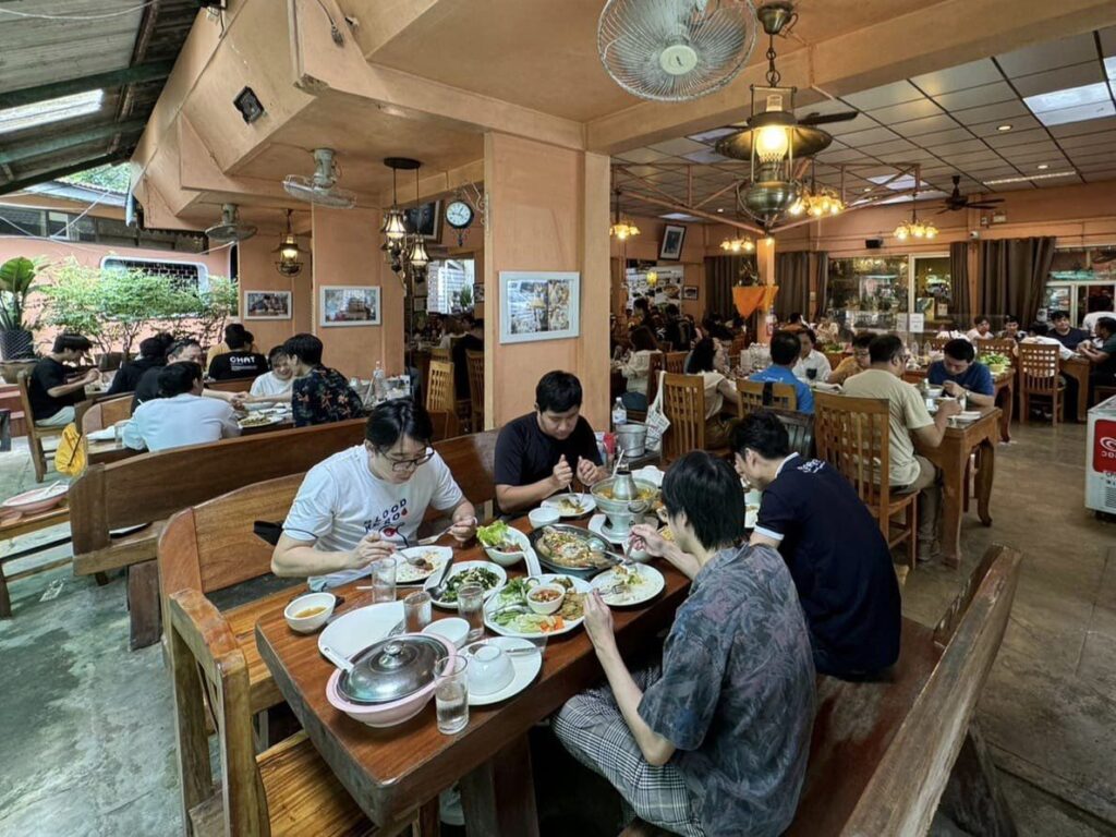 ร้านอาหารที่มีผู้คนพลุกพล่าน โดยมีผู้มารับประทานอาหารนั่งที่โต๊ะและม้านั่งไม้ กำลังรับประทานอาหาร  ท่องเที่ยวระนอง บรรยากาศเป็นกันเองด้วยพัดลมเพดานและการตกแต่งผนัง สร้างบรรยากาศที่มีชีวิตชีวาเหมาะสำหรับผู้ที่สำรวจระนอง