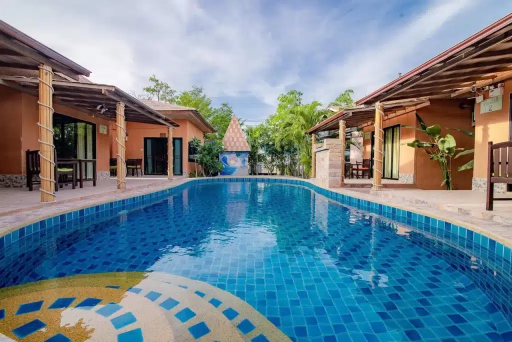 ฉากกลางแจ้งที่มีสระว่ายน้ำรูปสี่เหลี่ยมผืนผ้าที่ปูด้วยกระเบื้องสีฟ้า pool villa ใกล้กรุงเทพ โดยมีอาคารชั้นเดียว 2 หลังรายล้อมด้วยต้นไม้สีเขียวเป็นฉากหลัง ท้องฟ้ามีเมฆบางส่วน