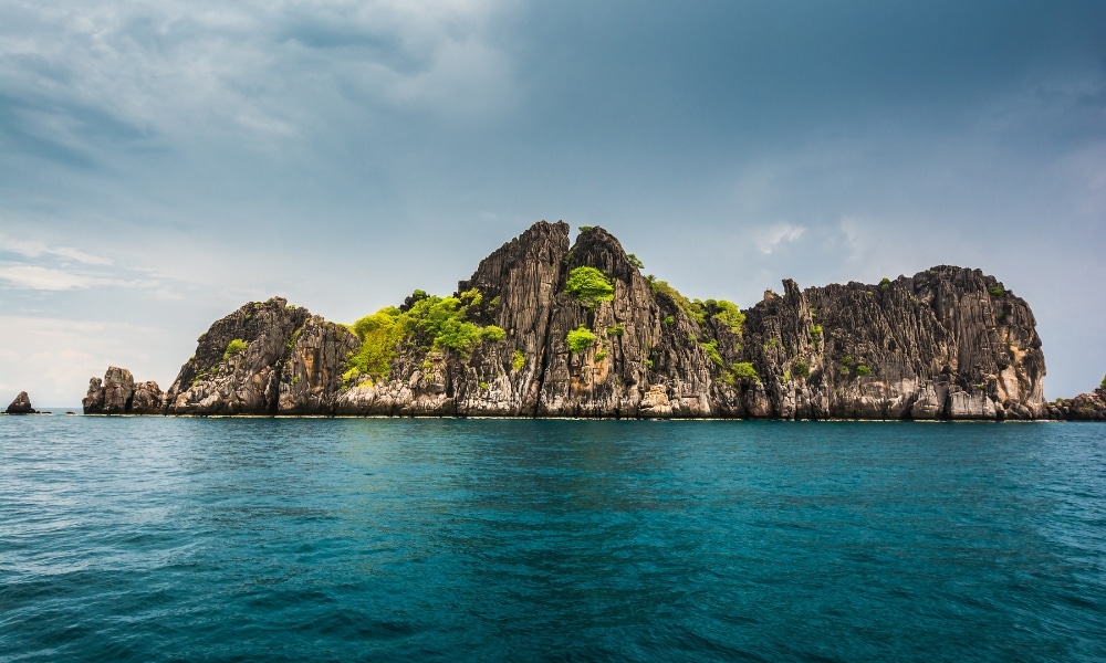 เกาะหินที่มีพืชพรรณสีเขียวล้อมรอบด้วยน้ำทะเลสีฟ้าอันสงบของภาคใต้ภายใต้ท้องฟ้าที่มีเมฆมาก ทะเลใต้สวยๆ