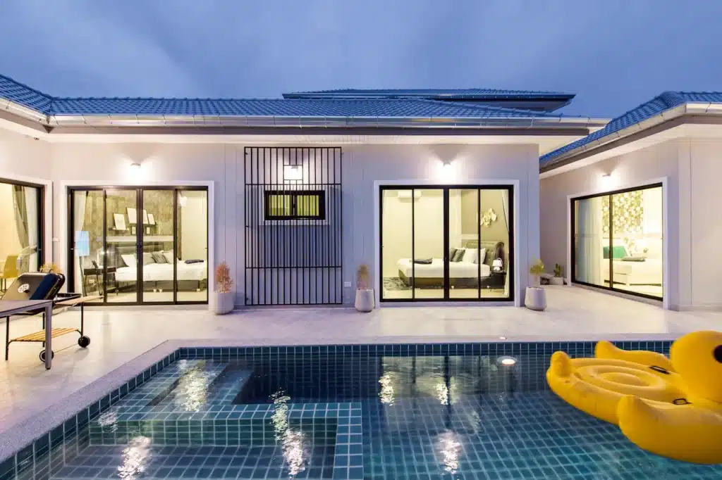 บ้านสไตล์โมเดิร์นที่มีหน้าต่างบาน pool villa ใกล้กรุงเทพ ใหญ่และประตูบานเลื่อน สระว่ายน้ำในตัวด้านหน้า และทุ่นลอยน้ำรูปเป็ดสีเหลือง แสงไฟภายนอกส่องสว่างบริเวณดังกล่าวในช่วงพลบค่ำ