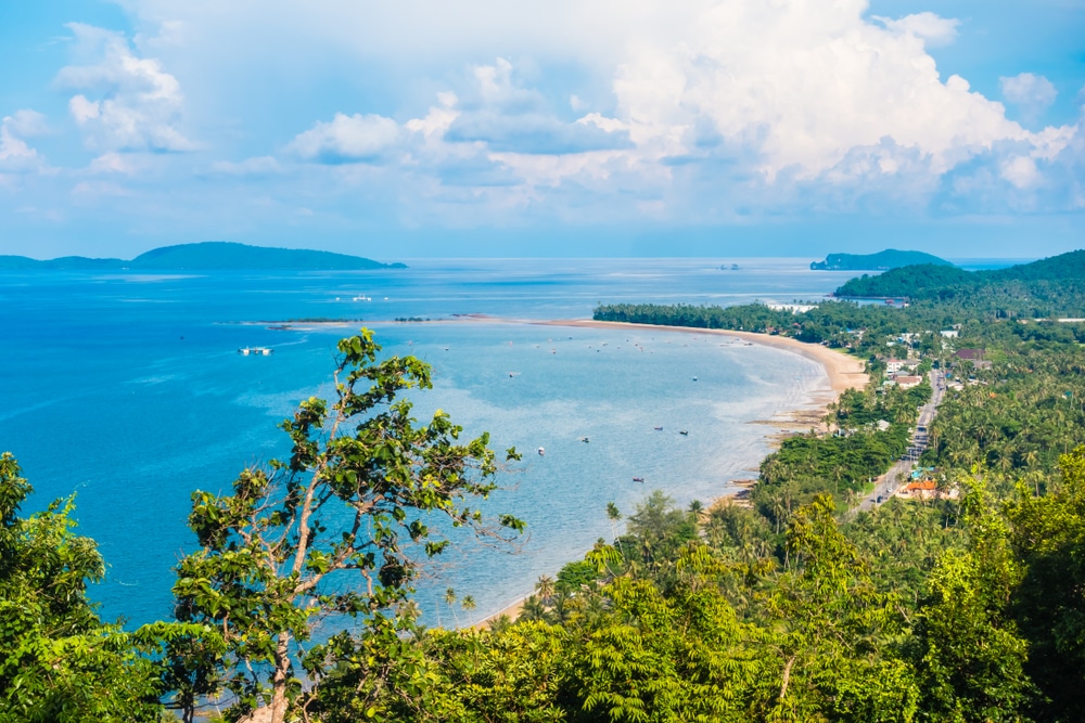 มุมมองทางอากาศของภูมิทัศน์ชายฝั่งเขตร้อนที่มีน้ำทะเลสีฟ้า หาดทราย พืชพรรณเขียวชอุ่ม และเรือที่กระจัดกระจายภายใต้ท้องฟ้าที่มีเมฆบางส่วน ชวนให้นึกถึง จุดชมวิวเขามัทรี ที่สวยงาม