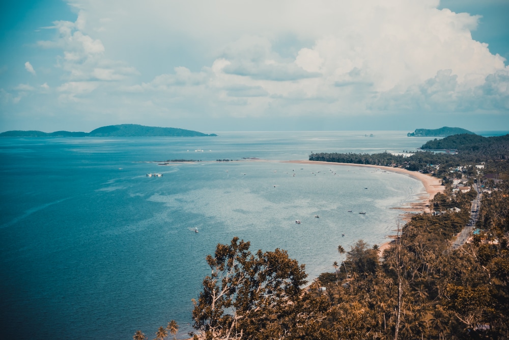 มุมมองทางอากาศของภูมิทัศน์ชายฝั่งทะเลที่มีหาดทรายโค้ง จุดชมวิวเขามัทรี น้ำทะเลสีฟ้าสงบ เรือลำเล็กหลายลำ และพืชพรรณเขียวชอุ่มภายใต้ท้องฟ้าที่มีเมฆบางส่วนที่จุดชมวิวเขามัทรี