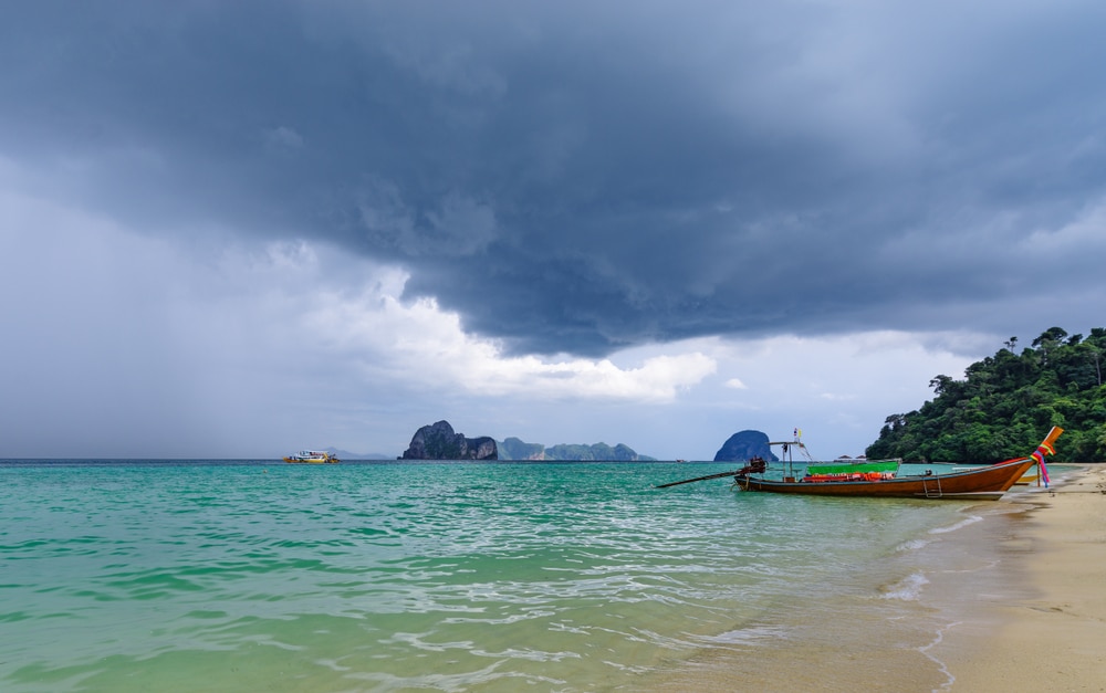 ชายหาดอันเงียบสงบพร้อมท้องฟ้ามีพายุ มีเรือแบบดั้งเดิมทอดสมออยู่ใกล้ชายฝั่งและมีเกาะสีเขียวชอุ่มเป็นฉากหลัง สะท้อนถึงที่เที่ยวทะเลใต้ได้อย่าง ทะเลใต้สวยๆ สมบูรณ์แบบ