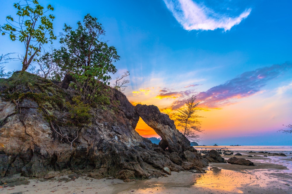 ฉากชายหาดหินยามพระอาทิตย์ตกดินพร้อมซุ้มหินธรรมชาติ ต้นไม้ และ ทะเลใต้สวยๆ น้ำลงเผยให้เห็นชายหาดทราย สีสันสดใสบนท้องฟ้าเปลี่ยนจากสีส้มเป็นสีน้ำเงิน สร้างฉากหลังที่สมบูรณ์แบบสำหรับการสำรวจ ที่เที่ยวทะเลใต้